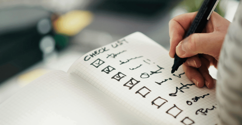 Zbliżenie na dłoń inżyniera jakości, który w notesie tworzy listę kontrolną (checklist) i odhacza niezbędne zwroty techniczne w języku angielskim, których się uczy.