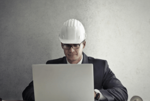 Inżynier jakości w białym kasku ochronnym siedzi przy biurku i pracuje na laptopie, przygotowując dokumentację i korzystając ze zwrotów technicznych w języku angielskim.