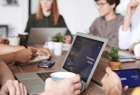 Zespół podczas spotkania jakościowego w biurze. Na ekranie laptopa widoczna jest agenda spotkania "Discussion Outline", a uczestnicy używają angielskich zwrotów do otwarcia dyskusji i podsumowania.