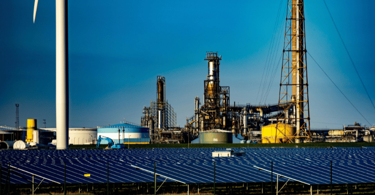 Transformacja energetyczna – turbina wiatrowa i farma fotowoltaiczna na tle rafinerii, symbolizujące przejście od paliw kopalnych do OZE.