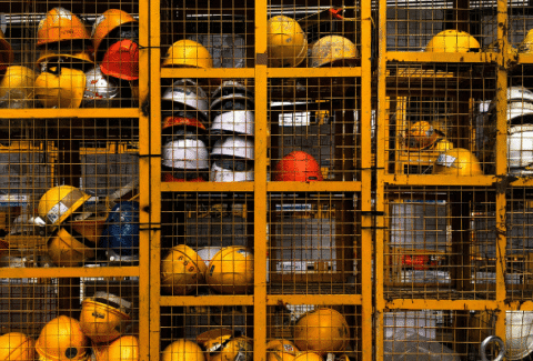 Żółte, białe i czerwone kaski ochronne ułożone w metalowych, kratowanych szafkach w zakładzie produkcyjnym. Grafika ilustruje znaczenie środków ochrony indywidualnej (PPE) w kulturze bezpieczeństwa w przemyśle.